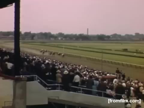 The still from 'Home Movies: Rochester/Niagara Falls 1948-1950' captures a lively horse racing event. A large crowd fills the stands, watching as several horses race down a verdant track. The scene is set against a pastel sky, evoking a nostalgic atmosphere typical of mid-20th century Americana. The image reflects the social and cultural activities of the time, showcasing leisure and community engagement.