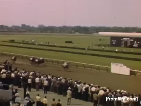The still from 'Home Movies: Rochester/Niagara Falls 1948-1950' depicts a vibrant scene at a horse racing event. Spectators are gathered along the racetrack, watching the competing horses, while a scoreboard displays the race details in the background. The overall atmosphere conveys excitement and community engagement typical of mid-20th-century American leisure activities. The colors and composition evoke a nostalgic feel of that era.