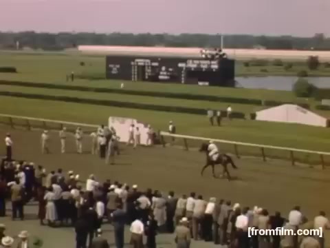 The still depicts a lively horse racing event, showcasing a group of jockeys on horseback nearing the finish line. A crowd of spectators, dressed in mid-20th century attire, watches eagerly from the stands by a racetrack. In the background, a large scoreboard displays race information, and a serene landscape with water is visible, indicative of the Rochester/Niagara Falls area during the late 1940s. The vibrant colors and composition capture the excitement of the event.