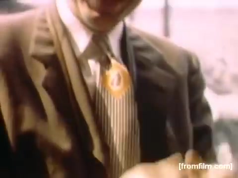 The still from 'Home Movies: Rochester/Niagara Falls 1948-1950' features a man smiling prominently in a suit, adorned with a button or decoration that suggests a celebratory context. The background suggests a casual outdoor setting, possibly during an event or gathering, depicting the joyful spirit of the time. The image reflects mid-20th century fashion and the social culture of that era.