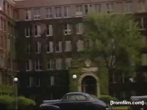 The still captures a mid-20th century scene featuring a large, brick building, likely an apartment or institutional structure, adorned with ivy. In the foreground, a vintage black car is parked. The setting suggests a suburban environment, reflective of Rochester or Niagara Falls in the late 1940s, showcasing the architectural style and urban landscape of that era.