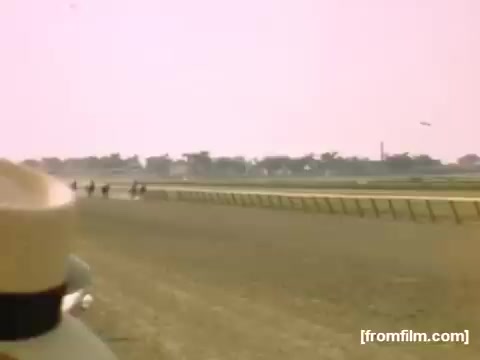 The still from 'Home Movies: Rochester/Niagara Falls 1948-1950' depicts a horse race at a racetrack, with riders in action along a sandy track. The foreground features a wide-brimmed hat, suggesting the perspective of a viewer or spectator. The background shows a blurred landscape, enhancing the dynamic feeling of motion, while the sky has a pinkish hue, adding a nostalgic atmosphere to the scene. The overall tone reflects the lively spirit of mid-20th century Americana.