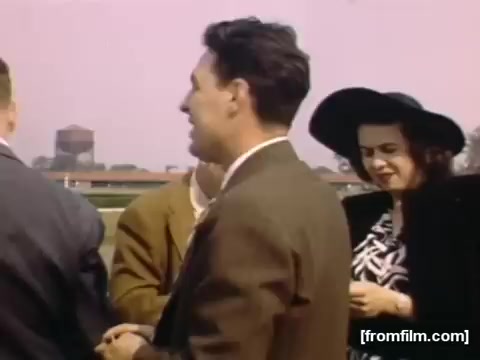 The still from 'Home Movies: Rochester/Niagara Falls 1948-1950' features a group of people outdoors, possibly at a social or community event. The individuals are dressed in mid-20th-century fashion, with a focus on a man in a suit and a woman in a large black hat and patterned dress. The background includes structures, hinting at an informal gathering atmosphere. The color palette suggests a vibrant, nostalgic view of life during that era.