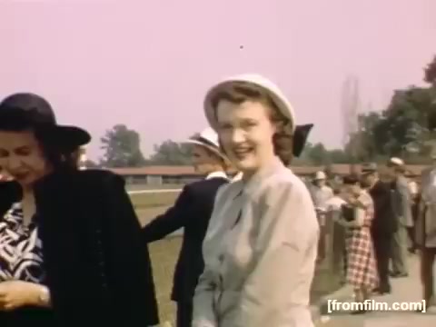 The still captures a moment from the film 'Home Movies: Rochester/Niagara Falls 1948-1950.' It features women in fashionable attire, one smiling broadly in a light-colored dress and hat, set against a backdrop of people gathered outdoors. The scene is characterized by a warm, nostalgic atmosphere, highlighting mid-20th-century social life. The color palette is soft, reflecting the aesthetic of the era.