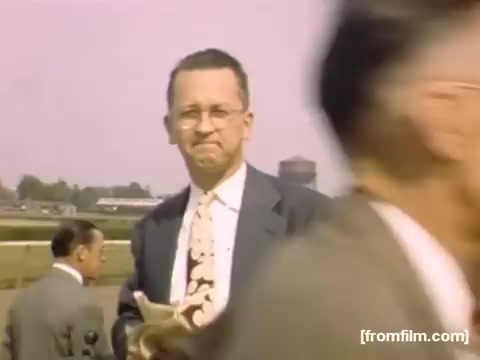 The still from 'Home Movies: Rochester/Niagara Falls 1948-1950' features a well-dressed man in a suit and tie, standing in a public space. He appears to be holding a piece of paper and looking slightly pensive. The background is blurred, suggesting movement or activity around him, with hints of other people and structures. The soft color palette and vintage style evoke a nostalgic atmosphere typical of the era.