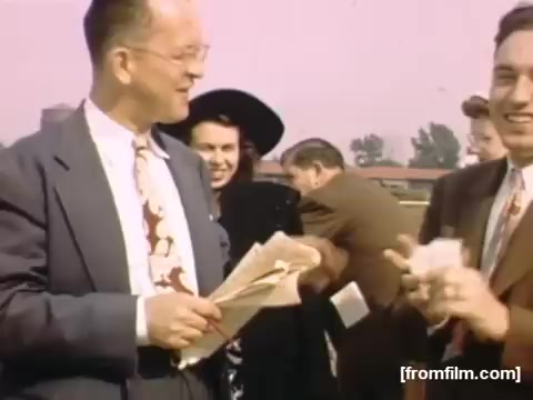 The 16mm still from 'Home Movies: Rochester/Niagara Falls 1948-1950' captures a lively outdoor scene featuring two men interacting, one holding papers and smiling, while another gestures playfully. In the background, several individuals, including a woman in a black hat, add to the social atmosphere. The pastel colors suggest a cheerful, nostalgic tone typical of post-war America.