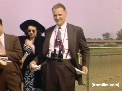 The still features a group of people at an outdoor event, likely a race or fair. A man in a suit stands smiling, with a camera attached to his waist. Behind him, a woman in a stylish outfit and sunglasses is partially obscured. The background shows a hint of a racetrack or open area, indicative of leisure activities popular in the late 1940s. The scene captures a moment of social interaction and leisurely enjoyment characteristic of the era.
