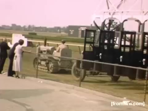The 16mm still from 'Home Movies: Rochester/Niagara Falls 1948-1950' depicts a scene at a racetrack. In the foreground, a small vintage tractor is parked next to a large black vehicle, possibly a starting gate for horse racing. A few people are leaning against a railing, observing the scene. The atmosphere suggests a lively event, with a backdrop of an expansive field and distant structures typical of mid-20th century America. The color palette is warm and slightly faded, characteristic of films from that era.