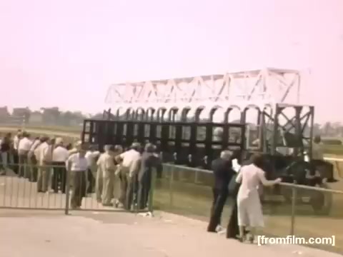 The still from 'Home Movies: Rochester/Niagara Falls 1948-1950' depicts a crowd gathered at a horse racing event. Attendees are lined up behind a barrier, watching a starting gate, likely waiting for the race to commence. The scene is set in a vibrant outdoor environment, characteristic of the era, with a pastel color palette that evokes a nostalgic feel.