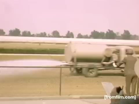 The still features a vintage scene from the late 1940s, depicting a truck labeled for spraying or irrigation in an agricultural or open-area setting. A person in a suit stands by, possibly observing or managing the operation. The color palette appears washed out, lending a nostalgic and historical quality typical of home movies from that era. The background is lined with trees or vegetation, contributing to the pastoral atmosphere.