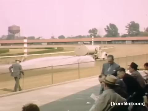 The still from 'Home Movies: Rochester/Niagara Falls 1948-1950' depicts a scene at a racetrack. It shows spectators seated in the foreground, observing a large water truck or grooming machine on the track, likely preparing the surface for a race. The environment is characterized by a vintage aesthetic, with vibrant colors and a clear sky, capturing the atmosphere of the time.