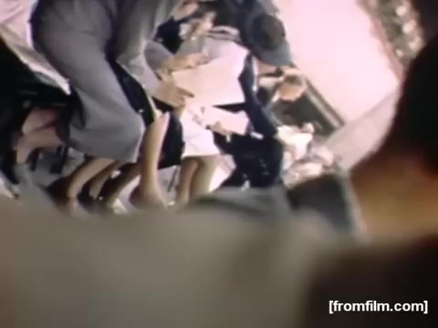 The still from 'Home Movies: Rochester/Niagara Falls 1948-1950' depicts a scene with several individuals seated, likely during an event. The focus is on their lower bodies and shoes, showcasing fashion from the late 1940s, with women in dresses and men in suits. The colors suggest a warm, nostalgic atmosphere, characteristic of home movie footage from that era. Overall, the image captures a moment of social gathering or leisure in a specific historical context.