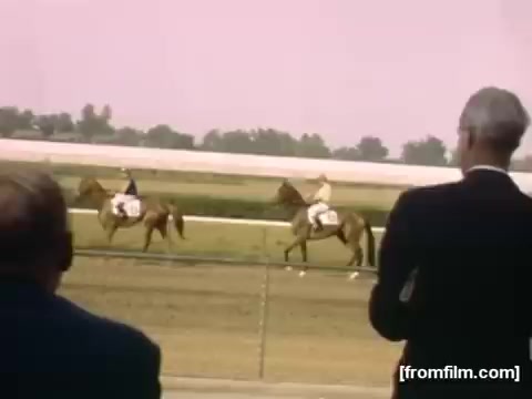 The still from 'Home Movies: Rochester/Niagara Falls 1948-1950' captures a horse racing scene, featuring two riders in colorful attire on horses. In the foreground, a couple of spectators in suits observe the race, highlighting the social atmosphere of the event. The background reveals a racetrack setting, with a lush landscape and a soft pink hue suggesting a warm day. This image reflects leisure activities popular in the post-war era.