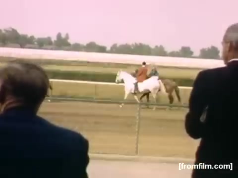 The still captures a scene from the film 'Home Movies: Rochester/Niagara Falls 1948-1950,' depicting two horses on a racecourse. One rider is on a white horse, while another rides a brown horse. The image features onlookers in formal attire watching intently, contributing to a nostalgic atmosphere that reflects horse racing culture in the late 1940s. The soft color palette adds a vintage feel to the moment.
