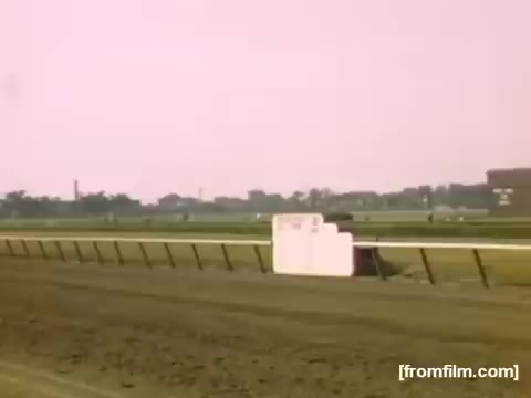 The still from 'Home Movies: Rochester/Niagara Falls 1948-1950' depicts a racetrack scene, likely during a horse race. The image features a dirt racetrack with a white barrier in the foreground and a blurred crowd or buildings in the background, set against a soft pastel sky. The overall aesthetic reflects the vintage nature of 16mm film from the late 1940s to early 1950s.