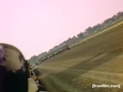 The still captures a dynamic scene from a horse race, showcasing a colorful, lively atmosphere typical of mid-20th century events. The image features blurred horses racing along a dirt track, with spectators visible in the foreground, emphasizing the excitement of the competition. The overall color palette contributes to a nostalgic, vibrant feel, representative of the period (1948-1950) and the cultural context of leisure activities in Rochester/Niagara Falls.