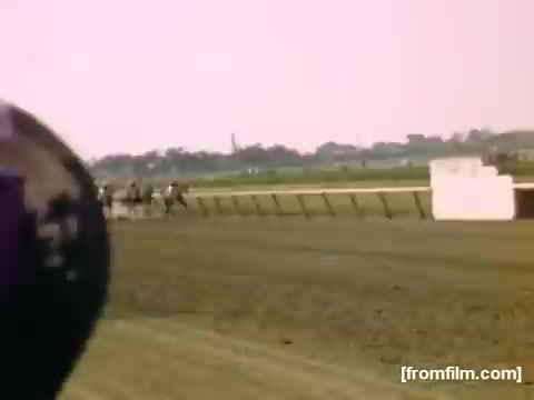 The still captures a moment from a horse race, likely at a racetrack, showcasing several horses racing across a dirt track. The vibrant colors reflect the mid-20th-century style, typical of home movie footage from 1948-1950. The focus is on the action of the race, with an out-of-focus object in the foreground, possibly a camera lens or a person's head, adding a personal touch to the scene. The background features a blurred landscape, hinting at the setting near Rochester or Niagara Falls.