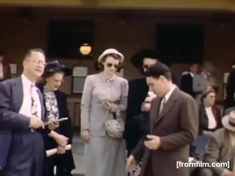 The still from 'Home Movies: Rochester/Niagara Falls 1948-1950' captures a group of well-dressed people in an outdoor setting, likely near a train station or similar venue. Several individuals are engaged in conversation, with one man in a suit appearing to present something to another, while a woman in a grey dress and hat observes. The scene exudes a vintage charm characteristic of the late 1940s, highlighting social interactions of the era.