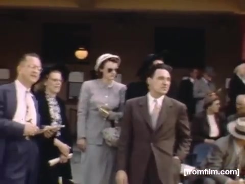 The still from 'Home Movies: Rochester/Niagara Falls 1948-1950' captures a gathering of people, likely in a social or public setting. A group of individuals, dressed in mid-20th century attire, are engaged in conversation or activity. The scene conveys a sense of community and nostalgia, reflecting the style and culture of the late 1940s. The color and composition suggest a casual yet significant occasion, possibly linked to local events in Rochester or Niagara Falls during that era.