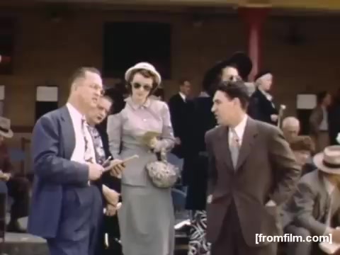 The still from 'Home Movies: Rochester/Niagara Falls 1948-1950' captures a curated social scene, likely at an outdoor event. Notable figures are dressed in formal attire, indicative of the late 1940s fashion. Two men engage in conversation, while a woman in a stylish gray outfit and hat holds a purse, suggesting an atmosphere of leisure and camaraderie. The background features additional attendees, hinting at a lively gathering, possibly a community event or a day at the races. The overall color and composition reflect the nostalgic feel of mid-20th century America.
