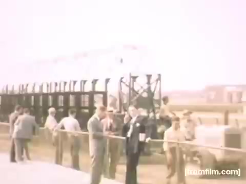 The still from 'Home Movies: Rochester/Niagara Falls 1948-1950' depicts a gathering of people, likely at a public event or site, featuring men in mid-20th-century attire, some with hats. They appear to be engaged in conversations, with a structure resembling scaffolding or a platform in the background. The overall atmosphere hints at a moment of communal significance or curiosity from this era. The color palette appears soft, typical of the film style from that period.