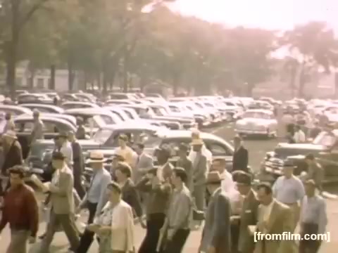 The still from 'Home Movies: Rochester/Niagara Falls 1948-1950' features a bustling scene from the late 1940s. A crowd of people walks past a large parking area filled with vintage cars, showcasing the fashion and social atmosphere of the time. The image captures a sense of community and leisure, likely at a public event or gathering. The warm color tones reflect the period's filmmaking style.