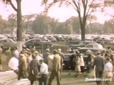 The still from **'Home Movies: Rochester/Niagara Falls 1948-1950'** captures a busy scene in a parking area filled with classic cars from the late 1940s. Groups of people, dressed in period attire, are walking together, suggesting a social or community event. The colors are warm and slightly faded, typical of 16mm film, conveying a nostalgic feel of the post-war era. The surrounding trees and distant vehicles create a sense of a lively atmosphere during that time.