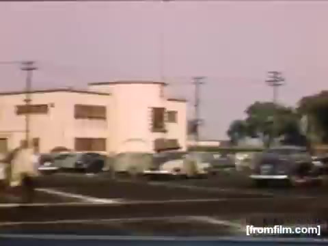 The still from 'Home Movies: Rochester/Niagara Falls 1948-1950' captures a bustling urban scene, showcasing a wide street lined with vintage cars. Prominent in the image is a large, white building with distinct architectural features, likely a commercial or public establishment of the time. The sky has a soft hue, suggesting early evening or late afternoon. People can be seen walking, adding a liveliness to the scene that reflects the era's everyday life.