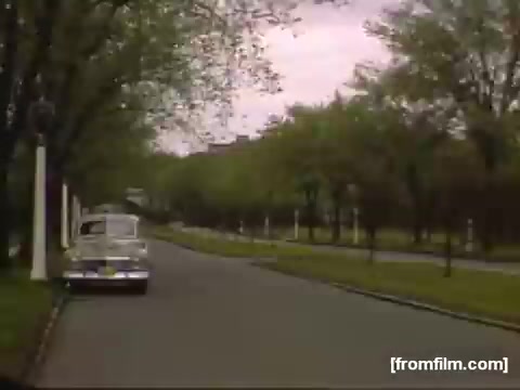 The still captures a serene street scene from the late 1940s, showcasing a tree-lined avenue with budding greenery. A vintage car is parked on the side, contributing to the nostalgic atmosphere. The soft pinkish hue of the sky hints at an overcast day, emphasizing the tranquil yet muted vibe typical of home movies from that era.