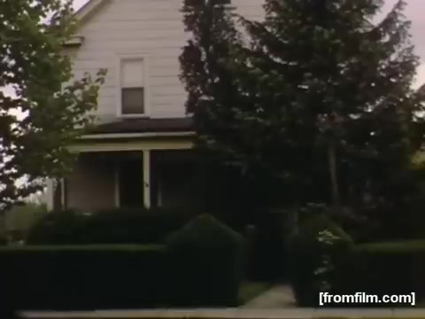 The still shows a residential house, characterized by its light-colored siding and a well-maintained front yard featuring neatly trimmed hedges and trees. The atmosphere reflects a suburban setting typical of Rochester or Niagara Falls during the late 1940s. The image captures a moment from the past, evoking nostalgia for home life in that era.