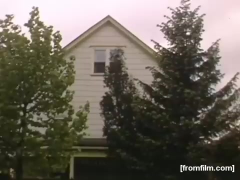 The still shows a close-up of a two-story house with a peaked roof, partially obscured by surrounding trees. The trees are lush and green, suggesting a warm season. The image reflects an intimate, nostalgic view typical of home movies, capturing a moment from the late 1940s to early 1950s in Rochester or Niagara Falls. The color palette appears soft and slightly muted, characteristic of 16mm film from that era.