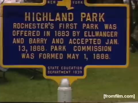 The still from 'Home Movies: Rochester/Niagara Falls 1948-1950' features a historical marker highlighting Highland Park, which was established as Rochester's first park in 1883. The sign details its acceptance date, the founding of the park commission, and includes a state education department designation from 1939. The image captures a moment of local history, illustrating community engagement with its natural spaces.