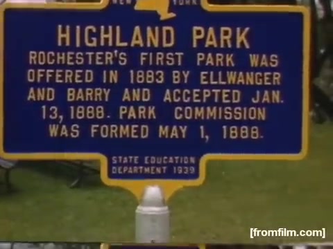 The still features a blue historical marker indicating 'Highland Park' in Rochester, New York. It provides details about the park's establishment, mentioning its creation in 1883 by Ellwanger and Barry, with the Park Commission forming on May 1, 1888. The sign emphasizes the park's significance as Rochester's first park. The date on the sign notes it was established in 1939.