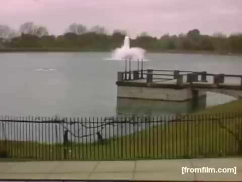 The still captures a serene scene by a lake, featuring a decorative fountain spraying water in the center. In the foreground, a wooden dock extends into the water, framed by a black wrought-iron fence. The surrounding landscape includes lush green grass, hinting at a well-maintained park environment. The sky is overcast, creating a calm and reflective mood typical of home movies from the late 1940s.
