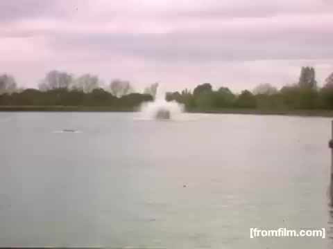The still from 'Home Movies: Rochester/Niagara Falls 1948-1950' shows a tranquil body of water, likely a lake or river, with a plume of water erupting in the center. The scene captures a moment of activity contrasted against the calm surroundings, with overcast skies and a tree-lined horizon, evoking a nostalgic, mid-20th-century atmosphere.