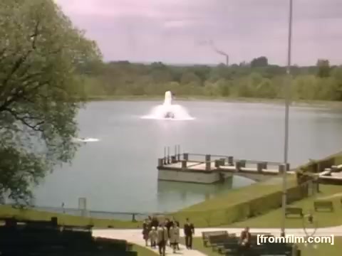 The still captures a serene scene from a park or lakeside area, featuring a fountain spraying water into a lake. In the foreground, a group of people strolls along a pathway, with benches visible for relaxation. Green trees frame the background, and a hint of industrial landscape can be seen in the distance, suggesting a juxtaposition of nature and urban life. The color tones reflect a nostalgic atmosphere typical of the late 1940s.