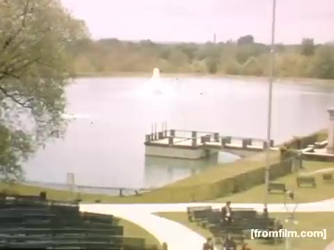 The still from 'Home Movies: Rochester/Niagara Falls 1948-1950' features a serene park scene centered around a lake. In the foreground, a neatly landscaped area includes benches, while a dock extends into the water. A fountain can be seen gently spraying water in the background, and lush greenery surrounds the lake, indicating a peaceful outdoor setting typical of mid-20th century leisure. The overall atmosphere is calm and picturesque, reflecting moments of everyday life during that era.