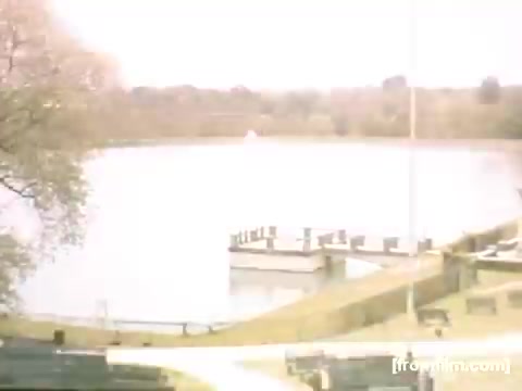 The still from 'Home Movies: Rochester/Niagara Falls 1948-1950' features a serene landscape with a body of water bordered by a grassy area and trees. There are wooden docks or piers extending into the water, suggesting a tranquil outdoor setting. The overall tone is nostalgic, capturing a peaceful moment from mid-20th century life in the Rochester/Niagara Falls area. The color appears soft and light, typical of 16mm film from that period.
