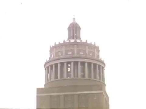 The still shows the top of a prominent building, likely the Times Square Building in Rochester, NY. The structure features a rounded, multi-tiered design with a tower topped by a spire. The image has a soft focus and light exposure, giving a nostalgic feel characteristic of 16mm film from the late 1940s to early 1950s. This context evokes a sense of mid-20th century Americana, reflecting the architectural style and urban landscape of that era.