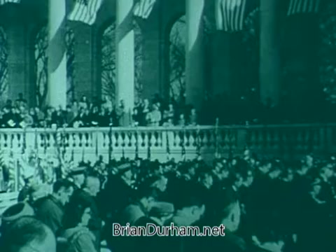 The still likely depicts a gathering for a Veterans Day public service announcement from the 1960s. It features a large audience, possibly outdoors, with veterans and spectators seated in rows, showing expressions of solemnity and reverence. American flags are visible in the background, indicating a memorial or commemorative atmosphere. The color tone appears to be muted, typical of the era's film style.
