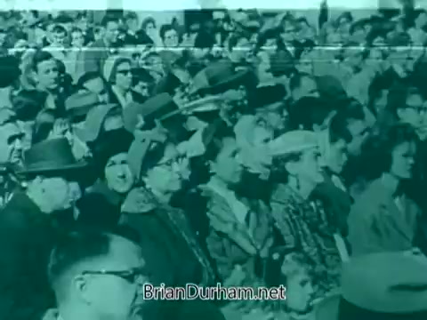 The still from the 1960s Veterans Day PSA captures a large crowd of individuals, predominantly wearing hats and varying styles of clothing typical of the era. They appear to be attentively listening or watching a presentation, likely in a public setting. The monochrome style gives it a vintage feel, emphasizing the gravity of the occasion. The focus appears to be on the collective response of the audience, highlighting a moment of reflection or commemoration.