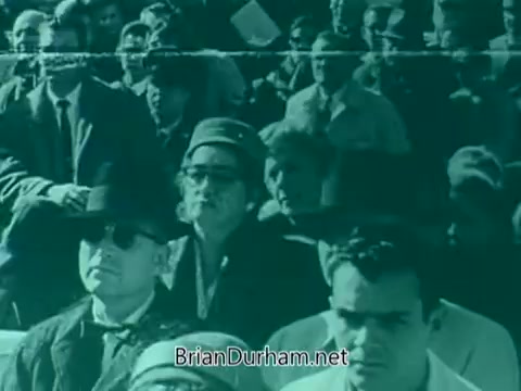 The still from the 'Veterans Day PSA - 1960's' features a crowd of people gathered for an event, likely a Veterans Day observance. The image is tinted in a blueish-green hue, giving it a vintage feel. Attendees are seen wearing hats and solemn expressions, suggesting a serious atmosphere. The focus on the audience indicates the importance of the event and the collective remembrance of veterans.