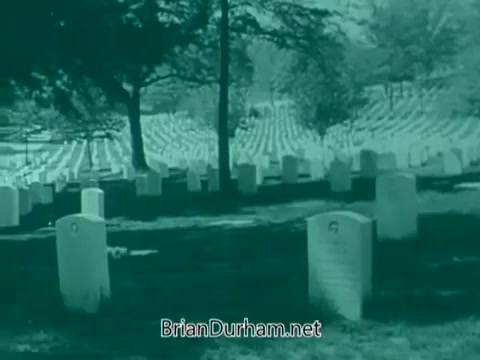 The still from the 'Veterans Day PSA - 1960s' depicts a serene cemetery scene, featuring rows of white gravestones arranged in a well-maintained grassy area. The environment is shaded by trees, creating a somber and respectful atmosphere, likely intended to honor and remember veterans. The color tone suggests an older film quality, reflecting the era's aesthetic.