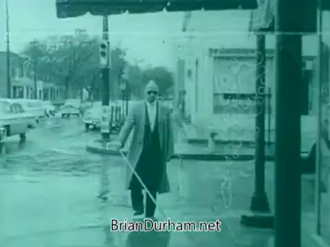 The still from the 'Veterans Day PSA - 1960's' depicts a man in a light-colored coat and sunglasses, walking with a white cane on a rainy street. The scene captures a somber, reflective mood, suggesting themes of resilience and support for veterans. The urban backdrop features vintage cars and storefronts, reinforcing the historical context of the 1960s. The overall aesthetic has a washed-out, teal tint characteristic of older film stock.