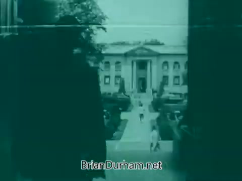 The 16mm still from the 'Veterans Day PSA - 1960's' features a view of a stately building framed by greenery. The image has a vintage aesthetic, indicative of the time period, and includes a group of people walking along a path in front of the building. The overall tone suggests a commemorative or reflective mood, appropriate for a Veterans Day message.