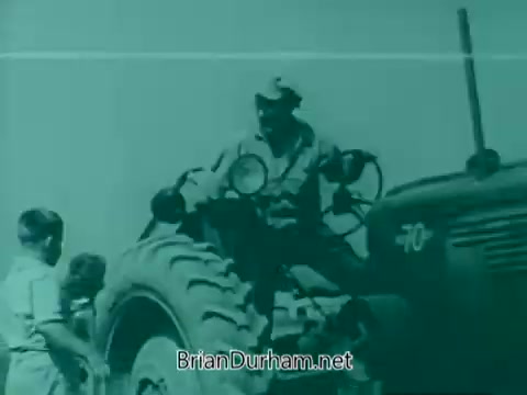 The still depicts a scene from the 1960s Veterans Day PSA, featuring a man on a tractor. He appears to be interacting with two individuals nearby. The image is characterized by a distinct teal tint, typical of older film technology. The tractor is prominently shown, illustrating a rural or agricultural setting, which may connect to themes of service and community.