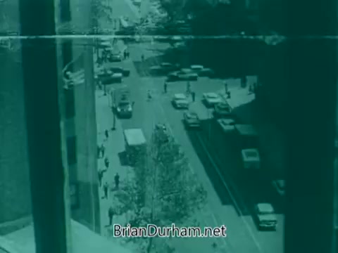 The 16mm still from the 'Veterans Day PSA - 1960's' depicts a busy urban street scene. It shows a diverse array of vehicles driving along the road and pedestrians walking on the sidewalks. The image has a greenish tint, suggesting an old film quality. There are buildings lining the street, capturing the atmosphere of a city in the 1960s.