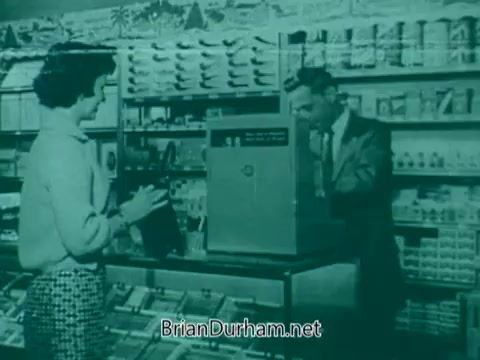 The still shows a scene set in a 1960s store, featuring a woman and a cashier at a counter. The woman, dressed in a light-colored blouse and patterned skirt, is holding a small black item, possibly a purse or bag. The cashier, a man in a suit, is engaging with her while standing behind a cash register. The backdrop is filled with various products and packaging, reflecting the retail environment of that era. The overall color tone has a vintage greenish hue, characteristic of the film style of that time.