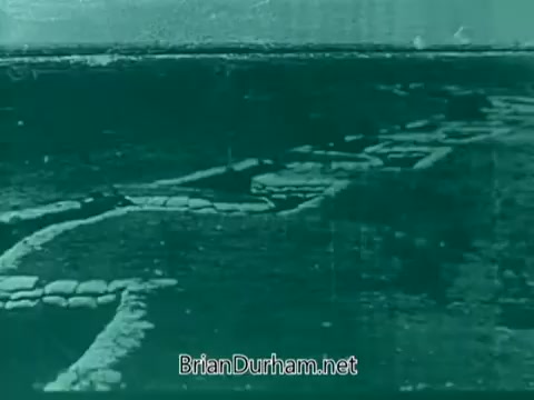 The still from the 1960s Veterans Day PSA depicts a landscape with what appears to be sandbags or fortifications laid out, possibly suggesting a military setting. The image has a muted green tint, creating a somber atmosphere appropriate for the context of honoring veterans. The emphasis seems to be on the landscape, evoking themes of sacrifice and remembrance.
