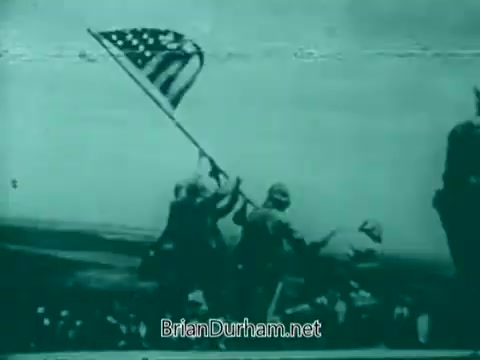 The 16mm still from the 'Veterans Day PSA - 1960's' depicts soldiers raising an American flag. The image conveys a sense of patriotism and camaraderie, likely aimed at honoring veterans. The visual style is characteristic of that era, possibly using monochrome or muted colors to emphasize the historical significance.